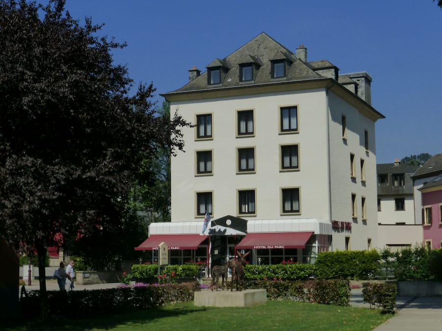 A multi-story hotel with a prominent red awning, a statue in front, and a well-maintained garden.