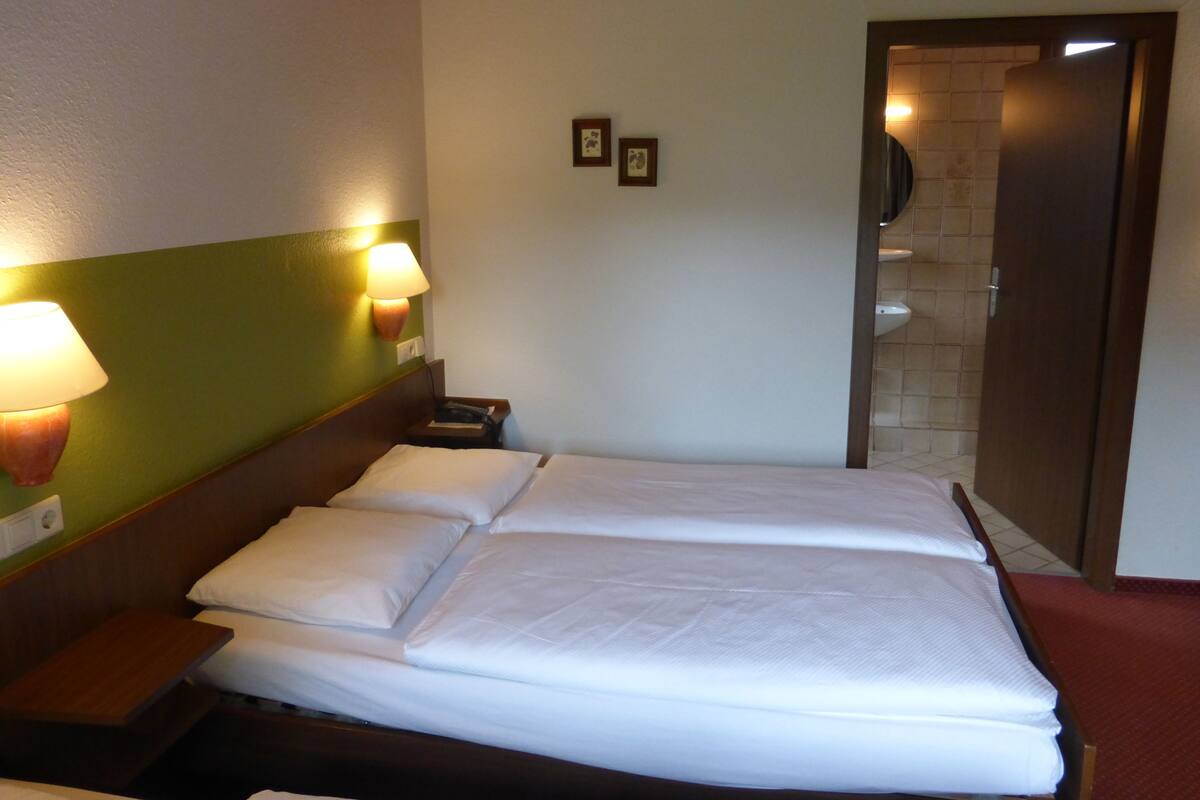 A hotel room with a large bed, two bedside tables, a nightstand, a wall-mounted lamp, and a door leading to a bathroom.