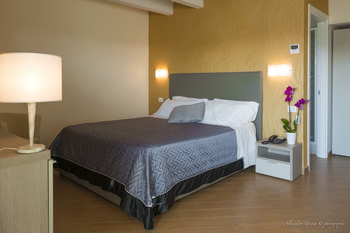 A neatly made bed with a grey quilt, a bedside lamp, a nightstand with a phone, and a potted orchid.