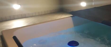 A hot tub with a blue light feature, surrounded by a tiled area with a low wall and a building in the background.
