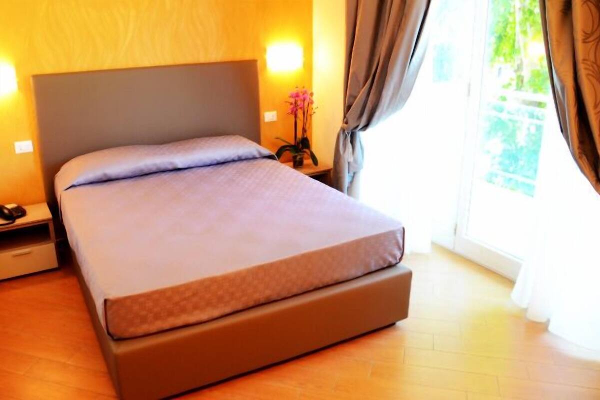 A neatly made bed in a room with a wooden floor, a nightstand with a lamp, and a window with curtains.