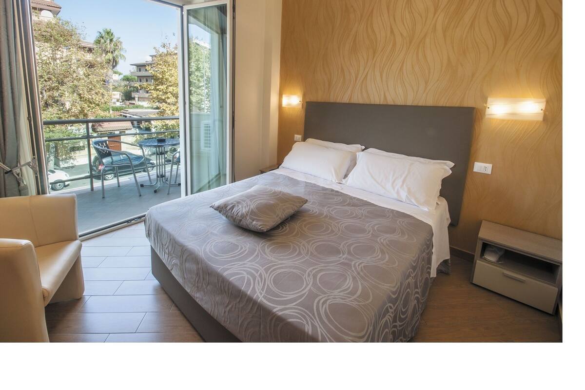 A bedroom with a bed, a chair, a balcony with a table and chairs, and a view of the outside.