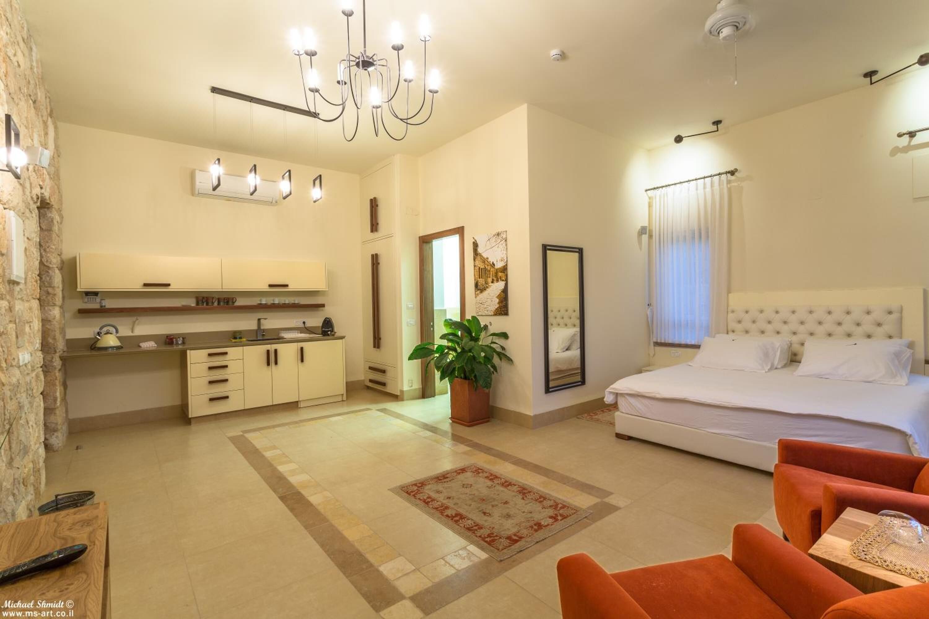 Une chambre d’hôtel moderne avec un grand lit, un coin salon séparé avec des fauteuils orange, un coin cuisine équipé d’un évier et de placards, et une plante en pot.
