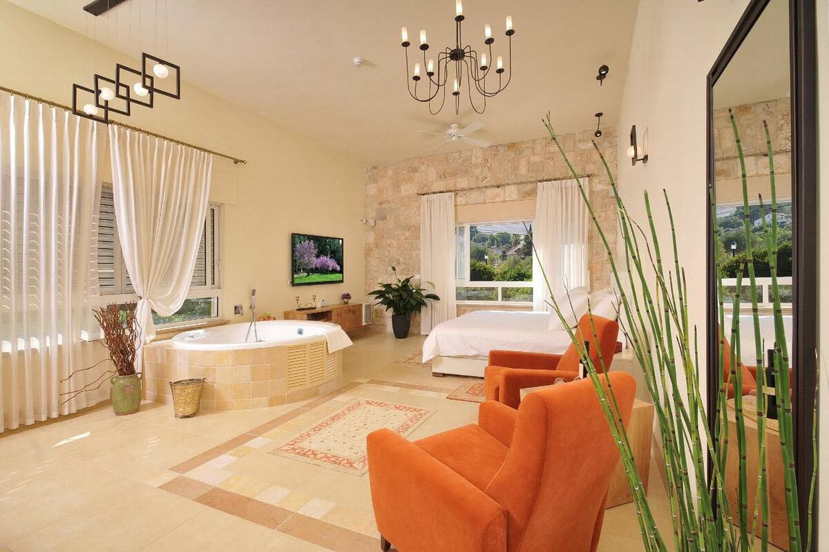 A spacious bedroom with a large bed, a sitting area with orange chairs, a bathtub, and a large window with curtains.