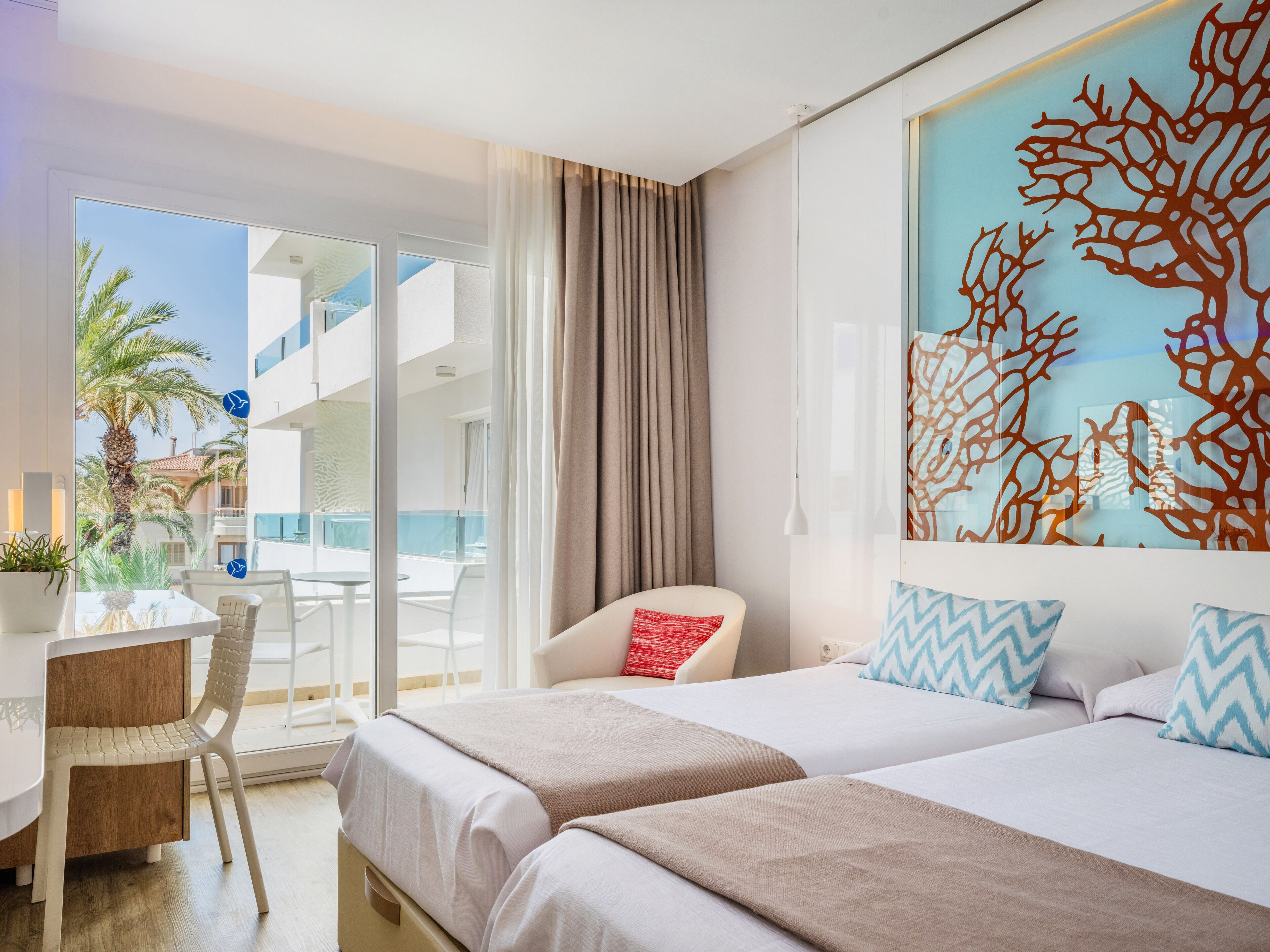 A hotel room with two beds, a desk, and a chair. There is a balcony with a view of palm trees and a pool.
