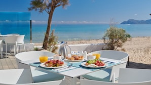 Una mesa de desayuno con platos de comida, una cafetera y un vaso de zumo de naranja en una terraza con vistas a la playa.