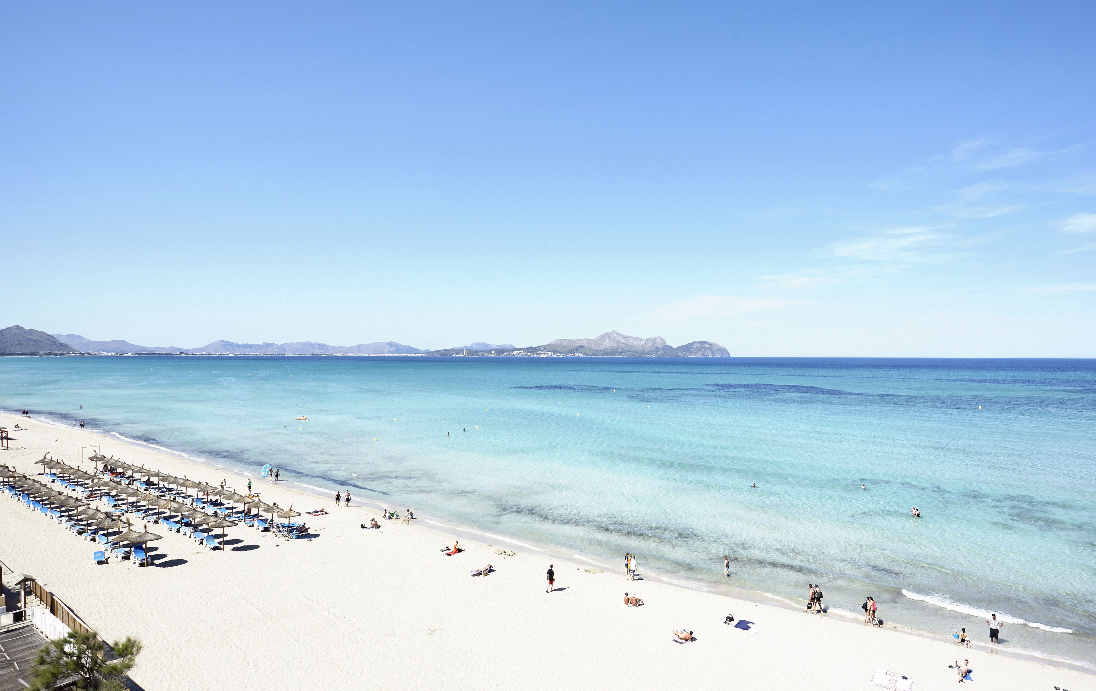 Una playa con agua turquesa cristalina, una orilla de arena y una hilera de sombrillas y tumbonas.