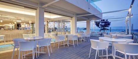 A modern outdoor dining area with white chairs and tables, a pool, and a view of the ocean.