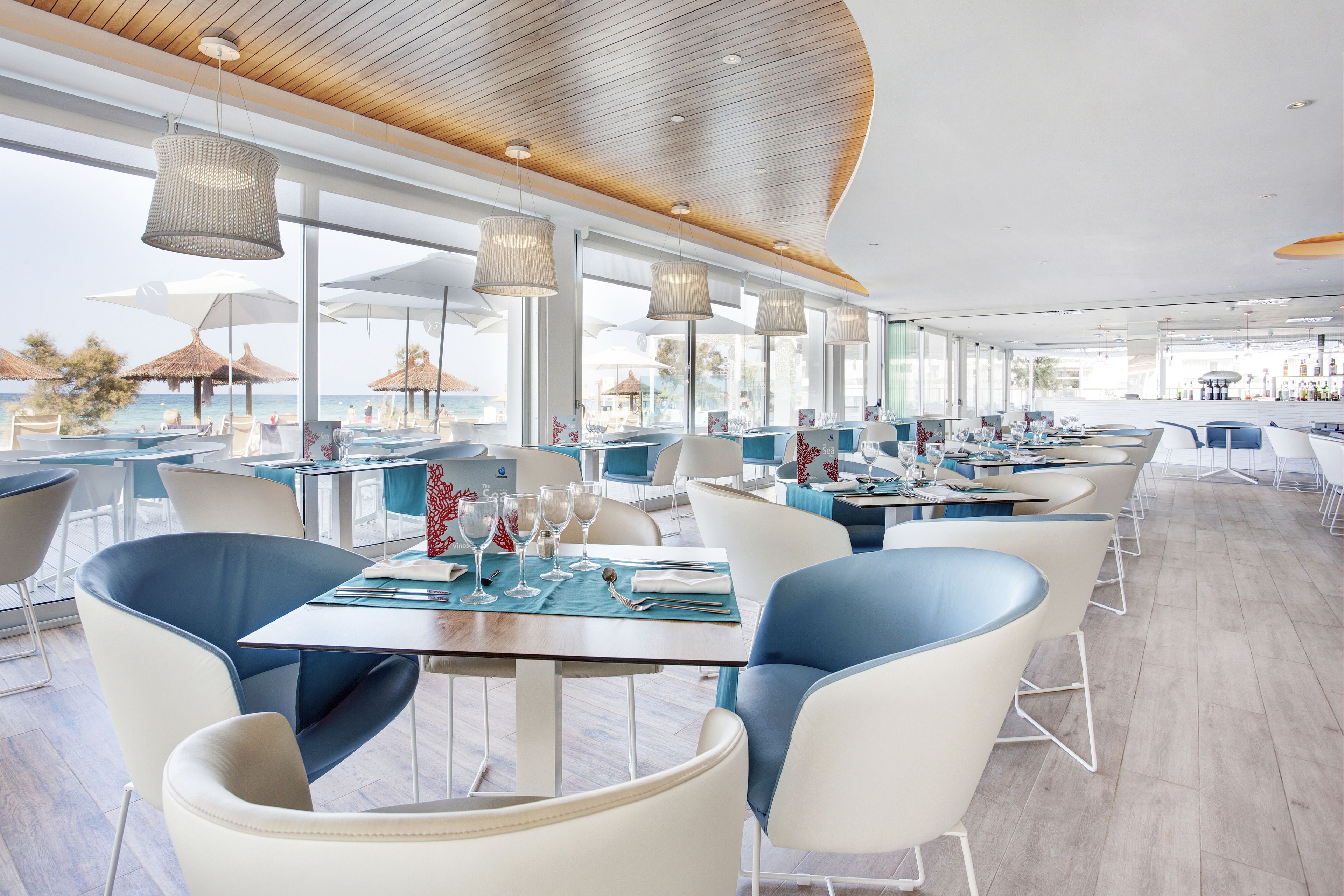 Een modern restaurant met uitzicht op zee, met witte en blauwe stoelen, houten tafels en hangende pendantlampen.