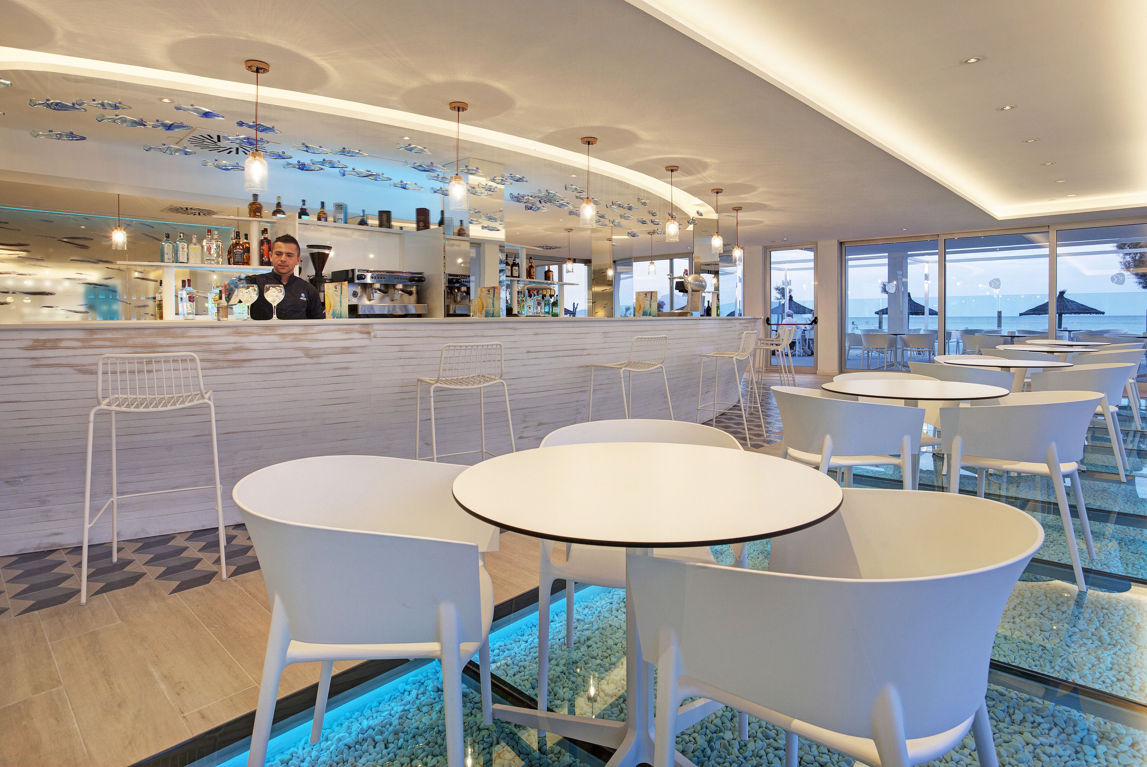 Un área de bar moderna con sillas y mesas blancas, una barra con botellas y vistas al océano a través de amplias ventanas.