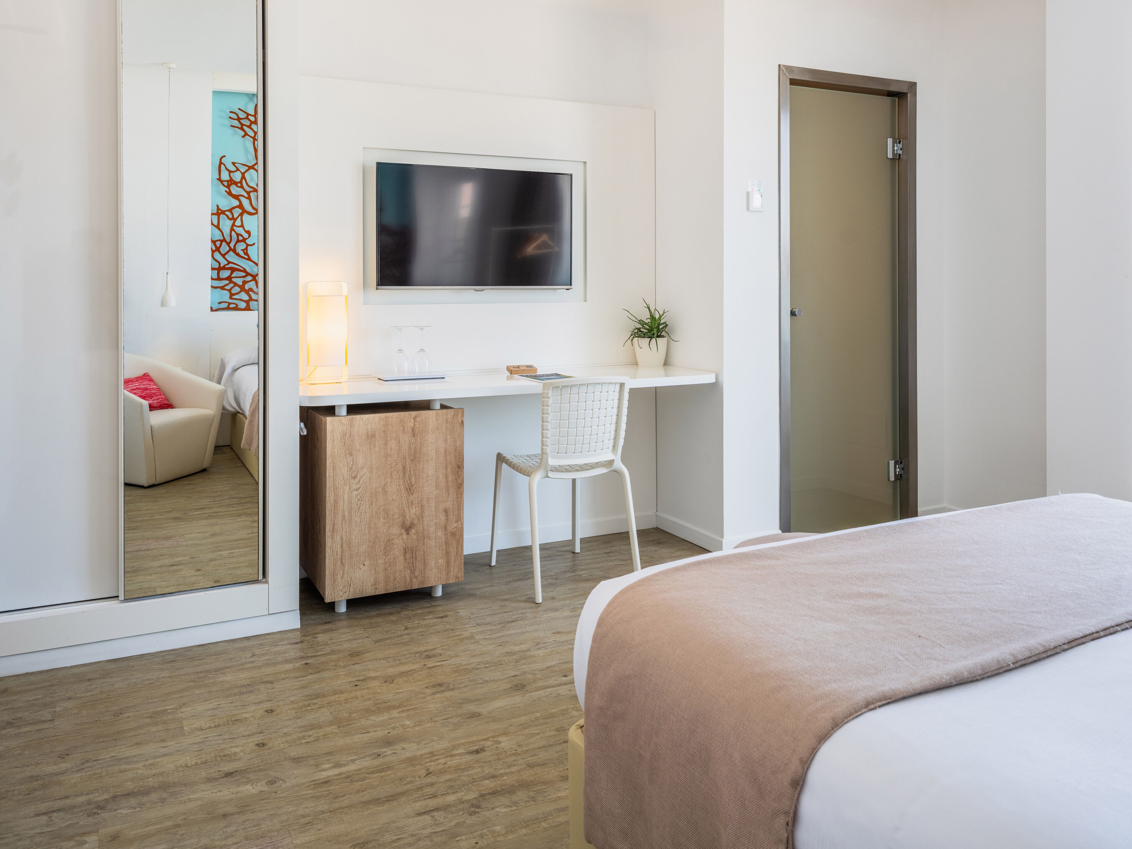 A modern hotel room with a bed, a desk with a computer, a chair, a small plant, and a mirror.
