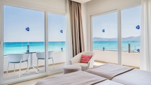 Habitación de hotel con un amplio ventanal corredero que da a un balcón con vistas al mar y a las montañas. Hay dos camas, una silla y una pequeña mesa con sillas en el balcón.