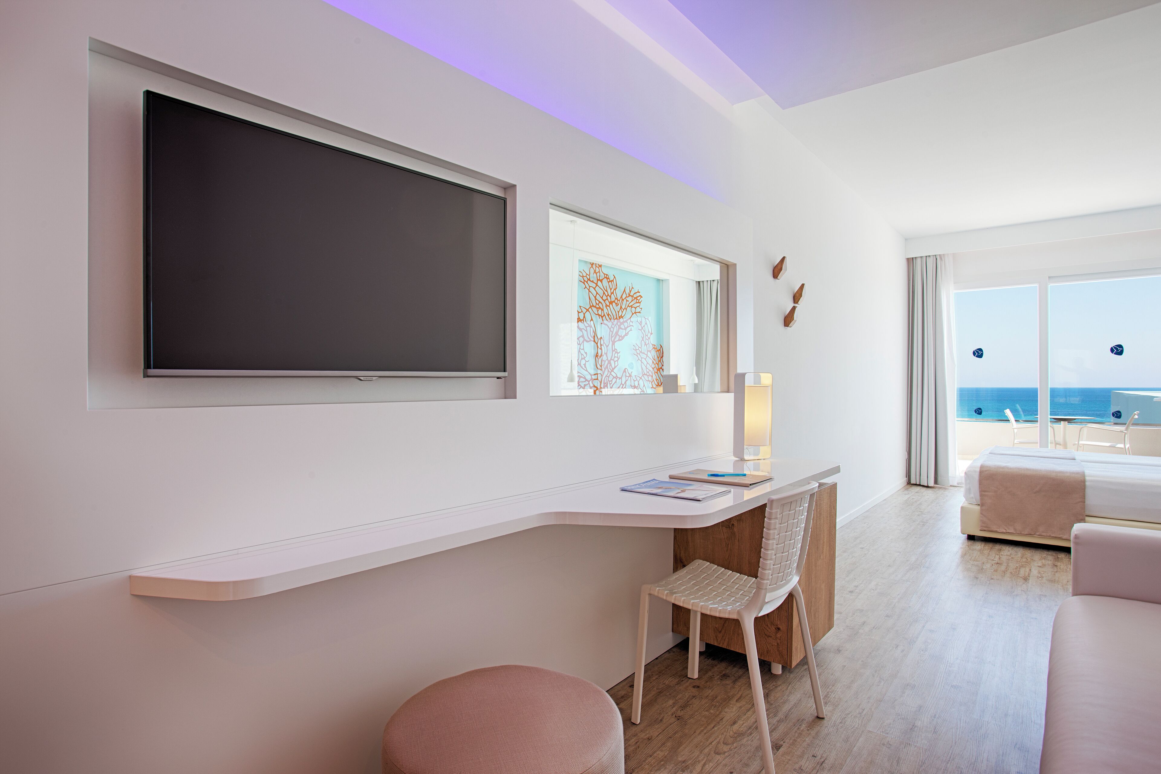 A modern hotel room with a large flat-screen TV, a desk with a chair, a bed, and a view of the sea.