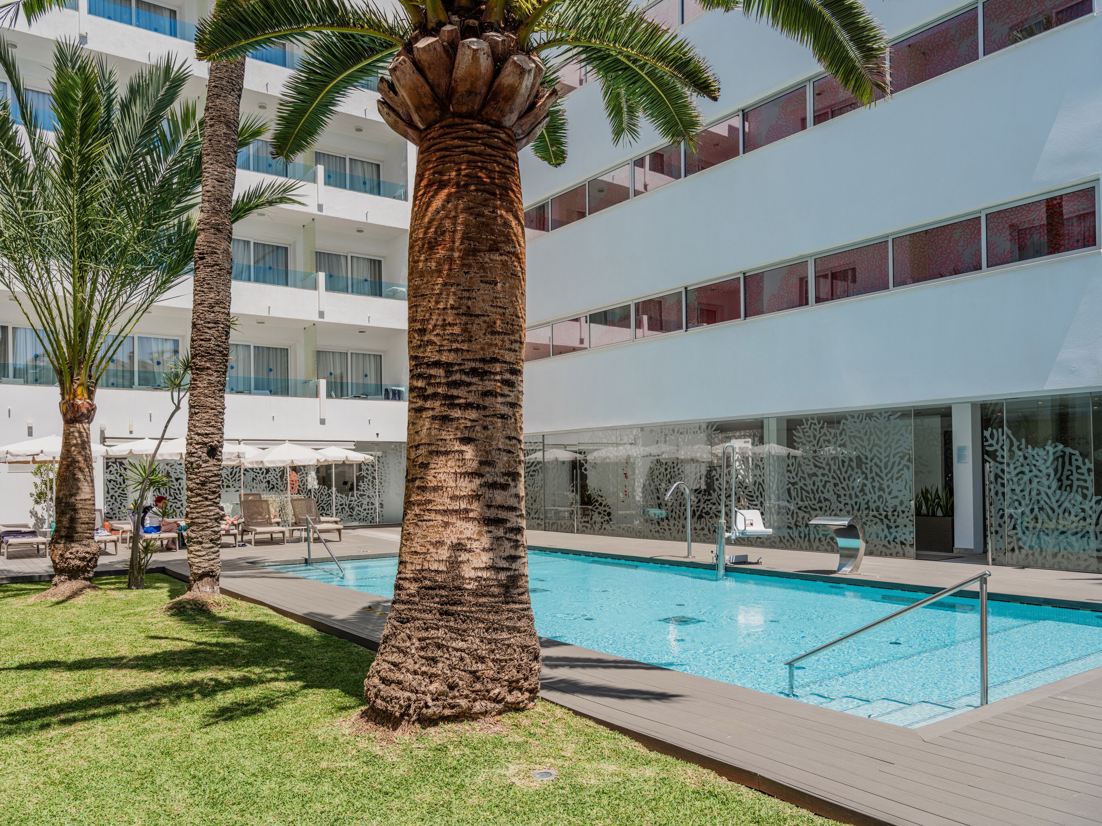 Un hotel moderno con piscina, palmeras y una zona de balcón con paredes de vidrio.
