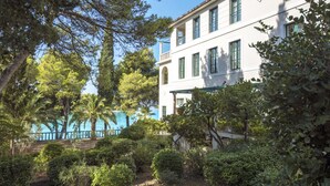 A white building with green shutters, surrounded by lush greenery and a clear blue sky.
