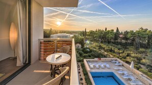 A balcony with a table and chairs, a pool with lounge chairs, and a view of a sunset over a landscape with trees and buildings.