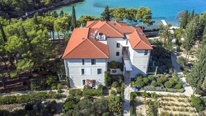 A two-story building with a red-tiled roof, surrounded by lush greenery and a clear blue sea.