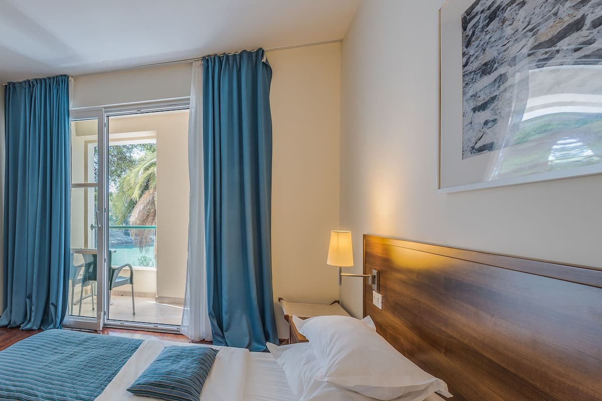 Hotellihuone, jossa on sänky, yöpöytä lampun kanssa, näkymä ulos ja seinällä oleva kehystetty kuva.