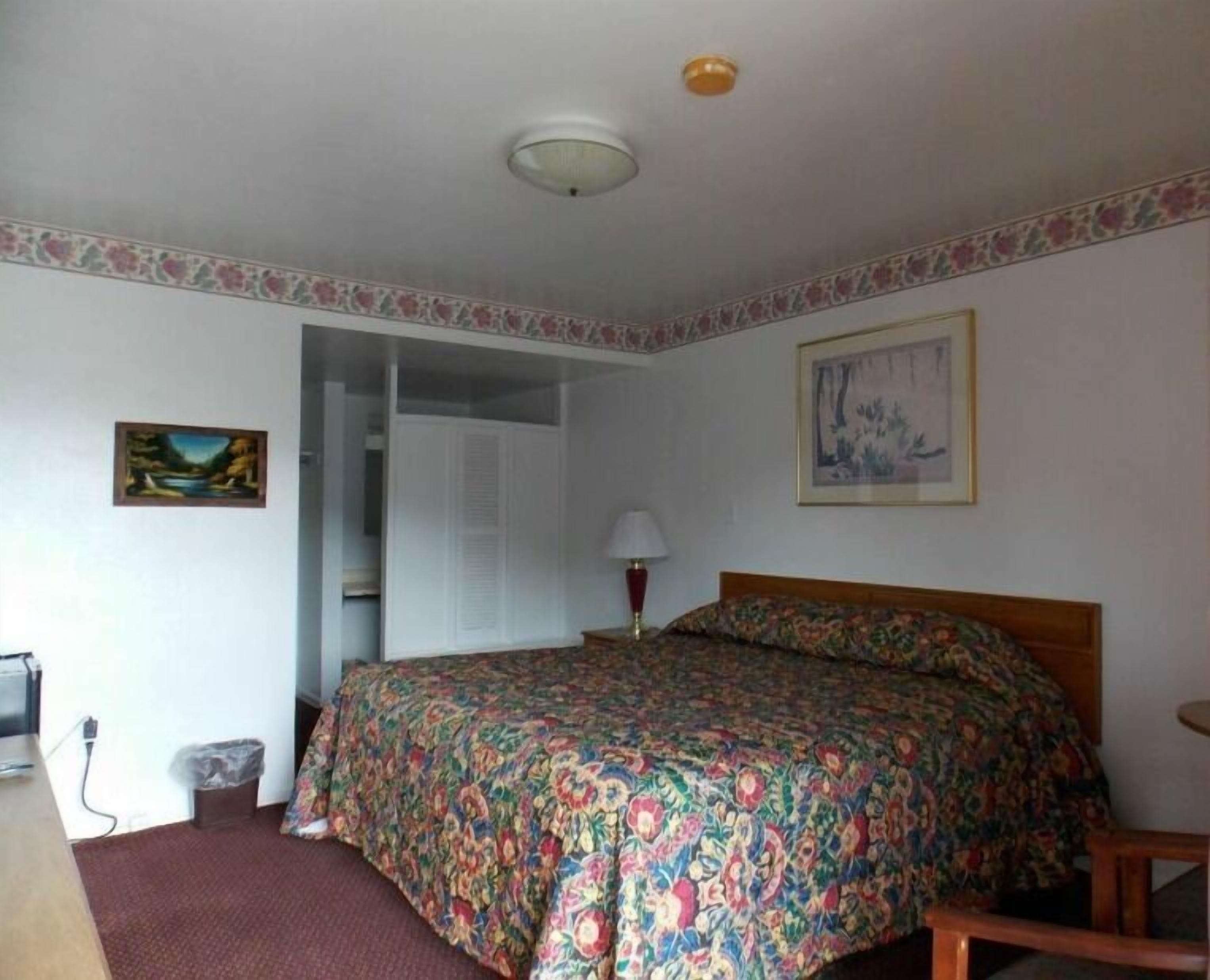 A bedroom with a bed, a nightstand, a lamp, a trash bin, and a door leading to another room.