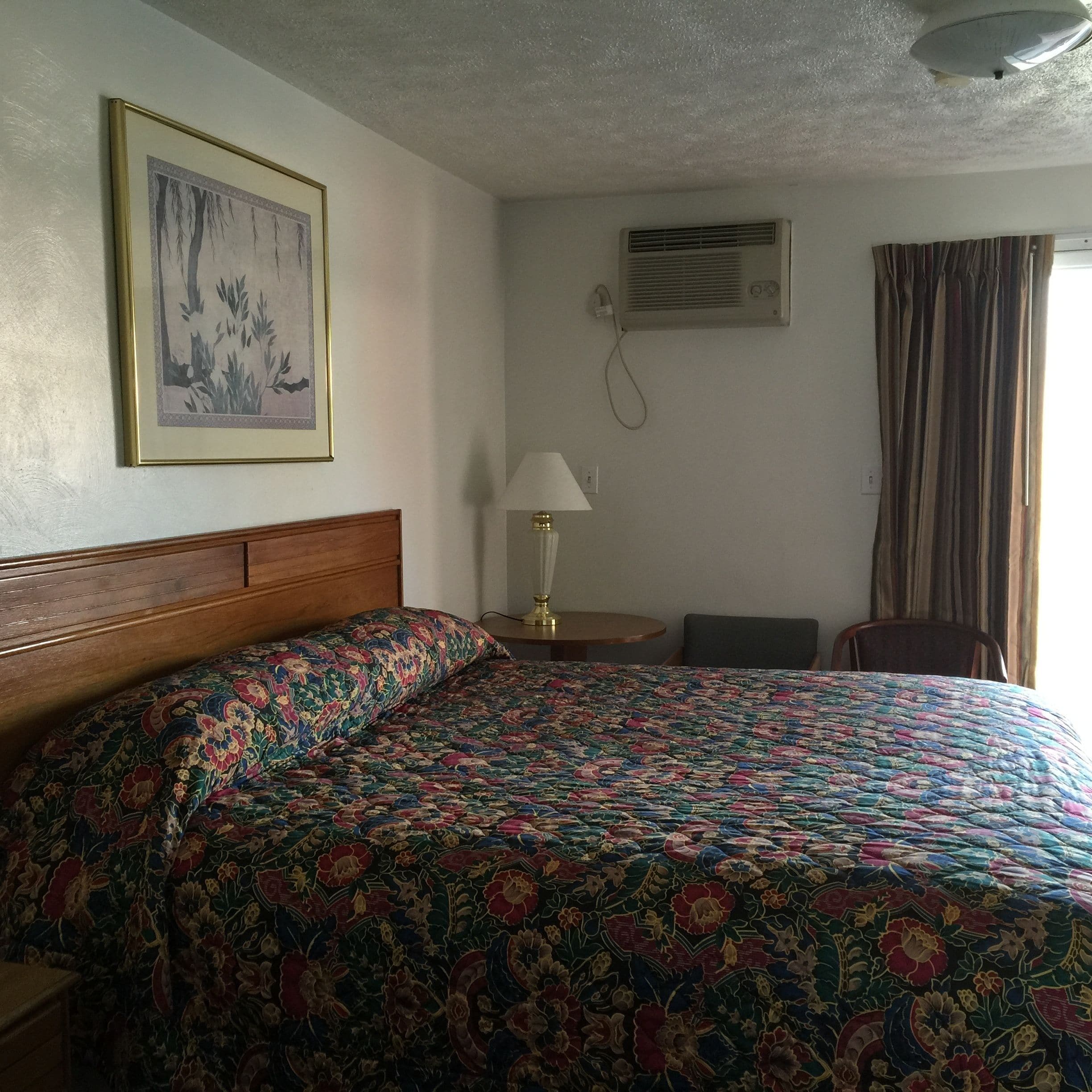 Ein Hotelzimmer mit hölzernem Kopfteil, einer farbenfrohen Blumenbettdecke, einem gerahmten Bild an der Wand, einer Klimaanlage, einer Lampe auf einem Nachttisch und einem Fenster mit Vorhängen.
