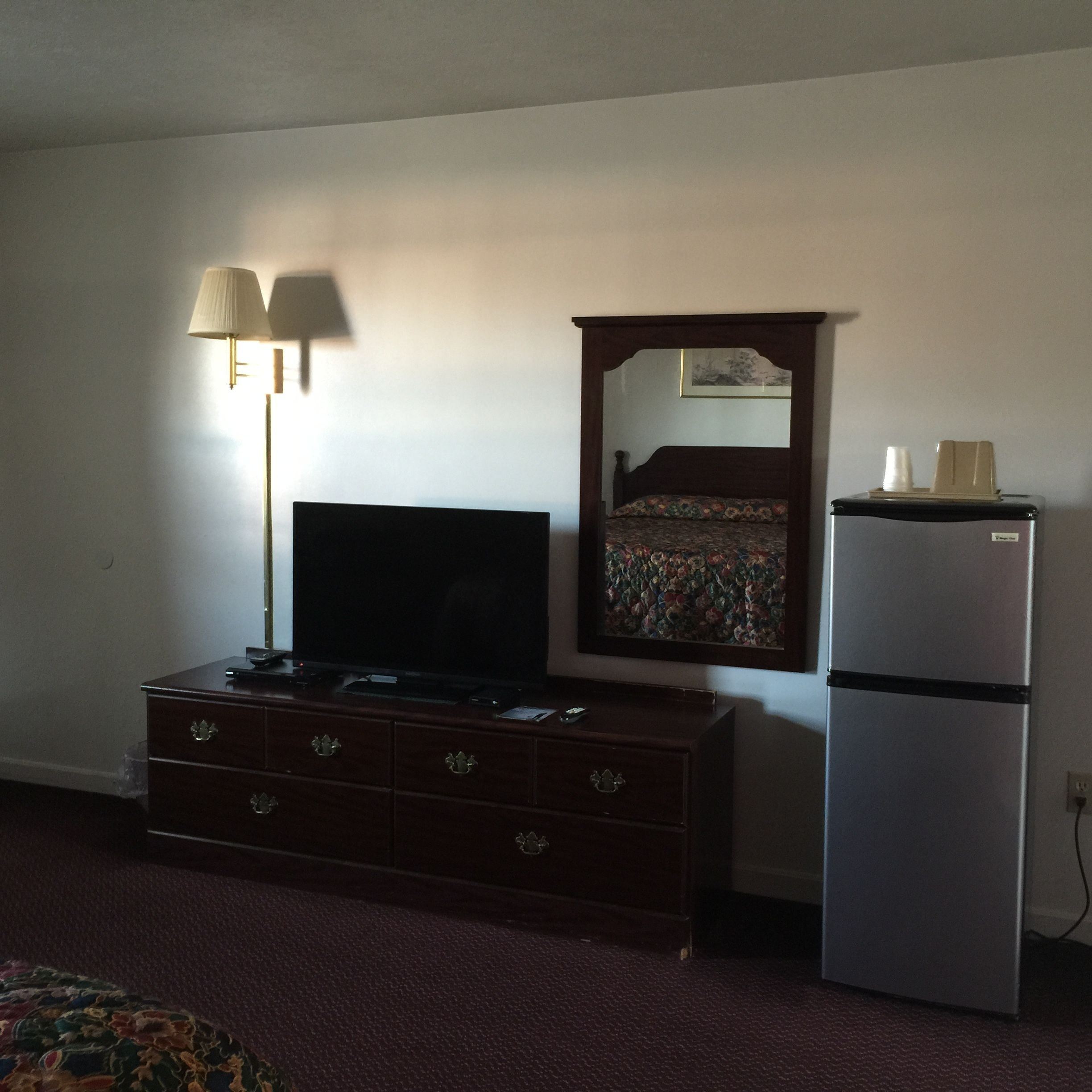 A hotel room with a dark wooden dresser, a flat-screen TV, a refrigerator, and a mirror with a floral bedspread reflected in it.