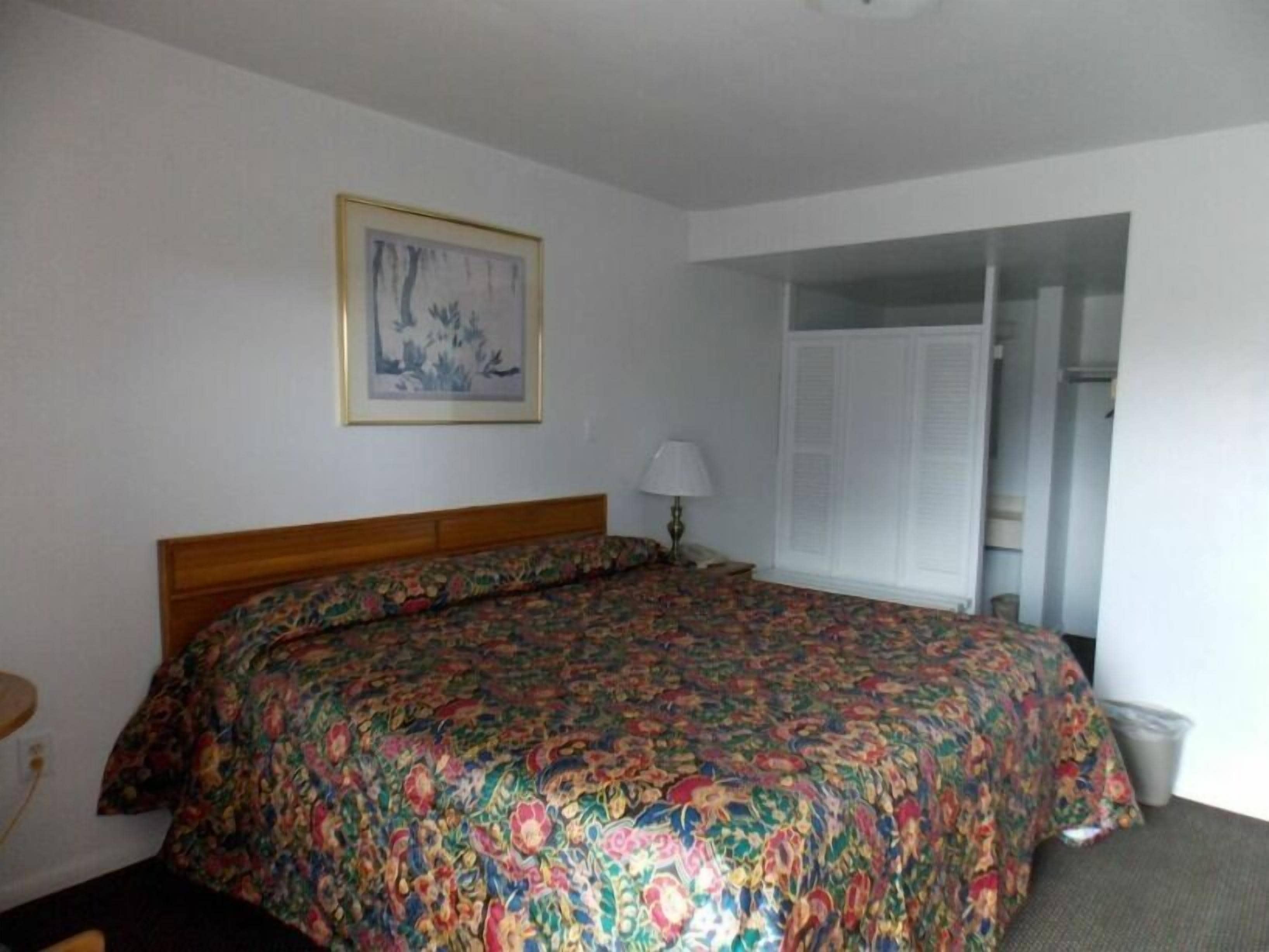 Quarto com cabeceira de madeira, colcha floral, luminária de cabeceira e um quadro na parede.