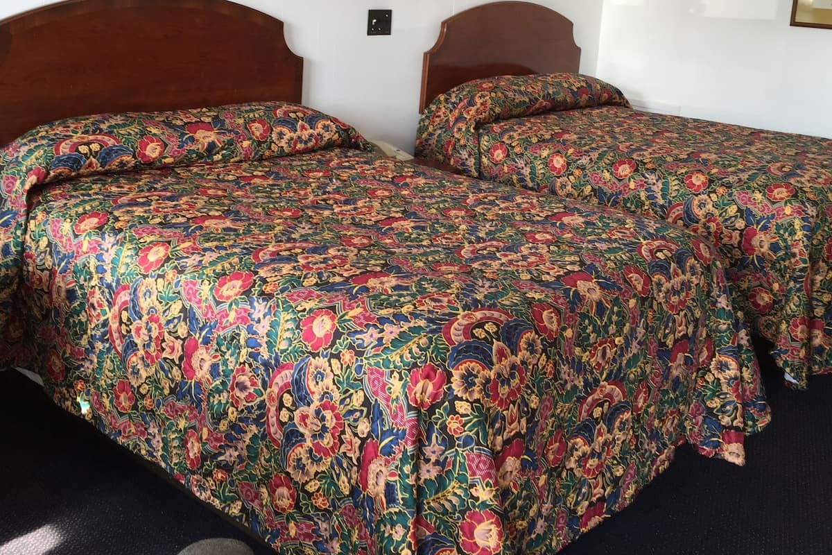 A hotel room with two wooden beds, each with a colorful floral bedspread. There are two wall-mounted lamps and a framed painting above the beds.