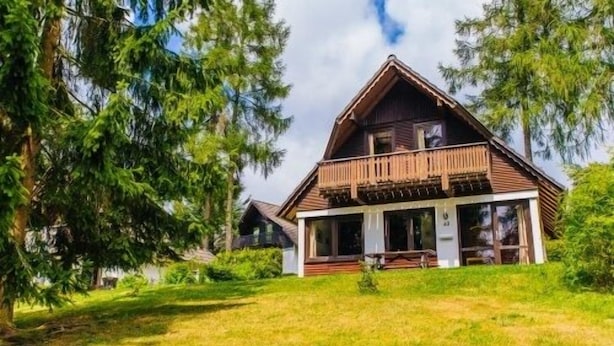 Een houten vakantiehuis met twee verdiepingen en een balkon, omringd door bomen en een gazon.