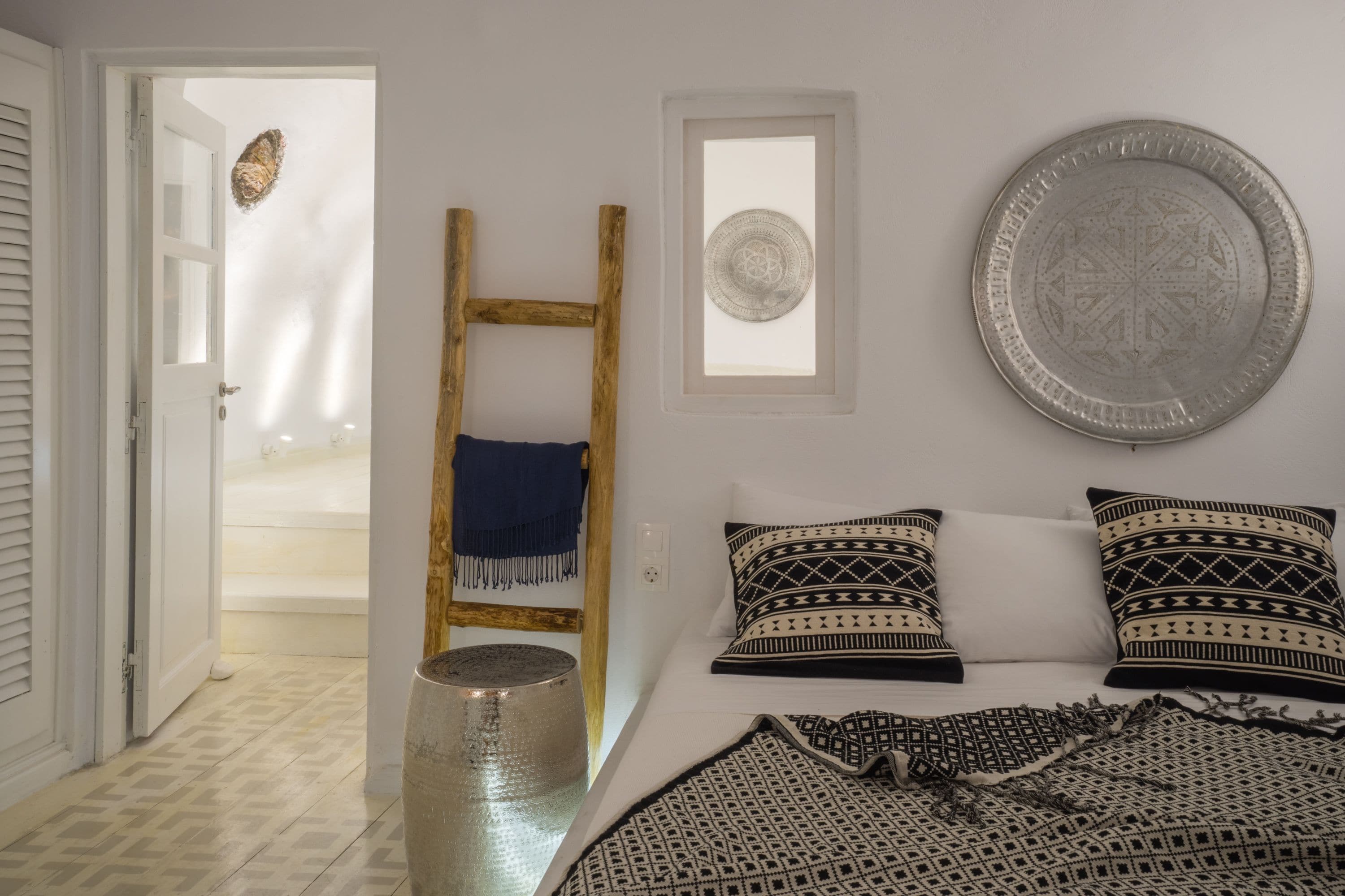 A bedroom with a bed, patterned pillows, a wooden ladder, a decorative plate, and a small window.