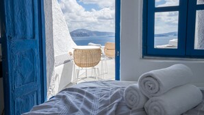Una habitación con una cama, dos sillones de mimbre, una ventana con vista al mar y las montañas, y una puerta azul.