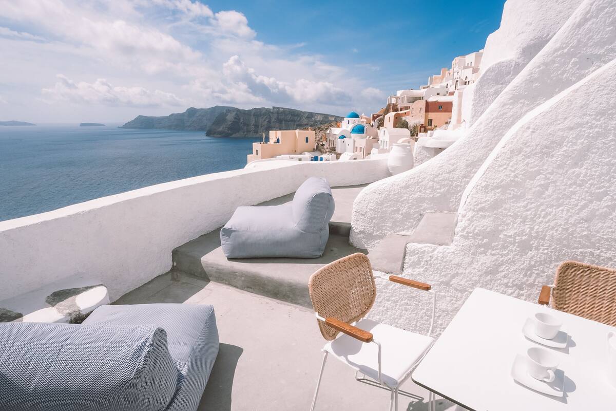 Una terraza frente al mar con edificios de paredes blancas, vista al océano y mobiliario exterior.