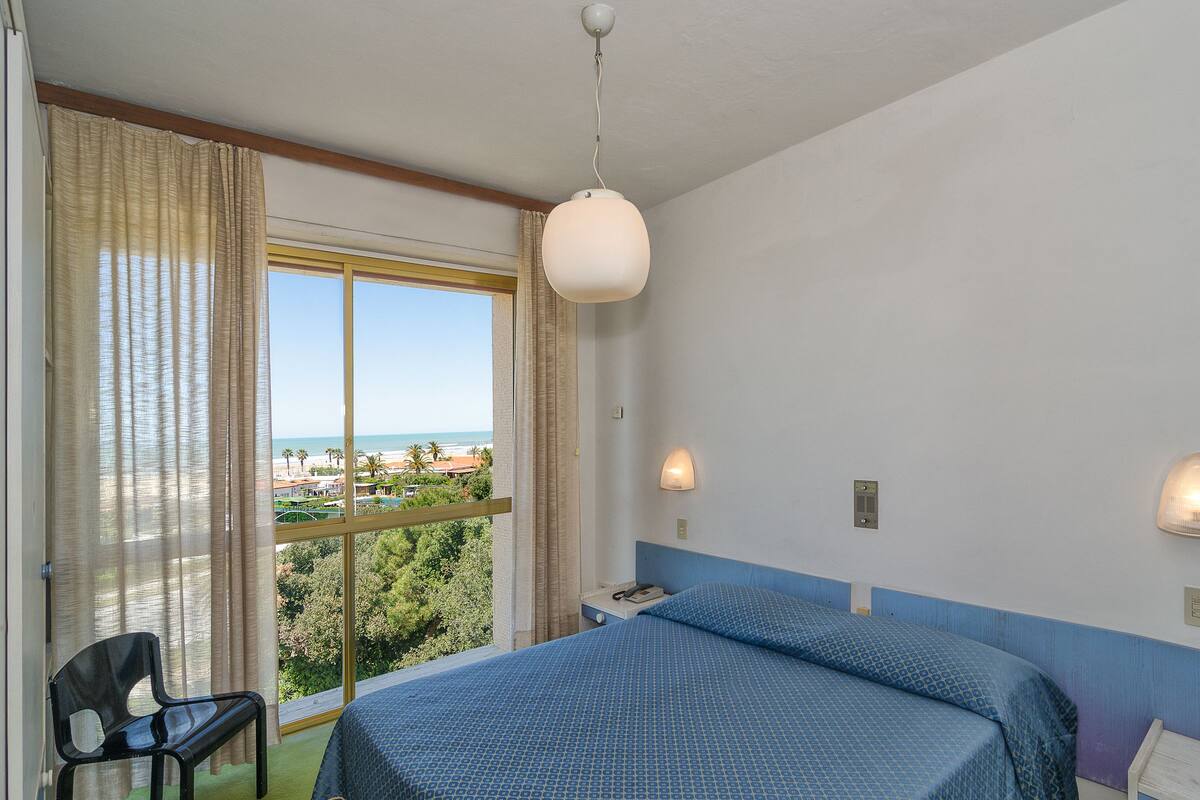 Quarto de hotel com cama, uma cadeira, mesa de cabeceira, luminária e vista para a praia.