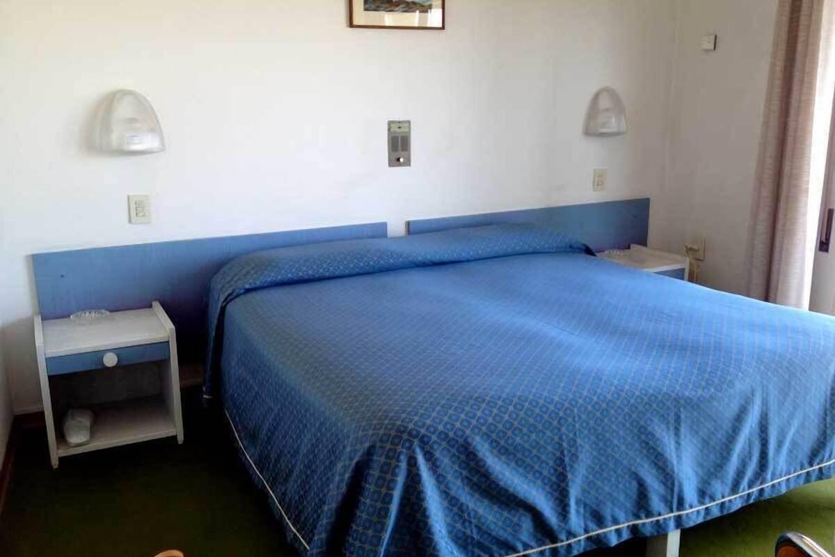 Quarto de hotel com uma cama de casal, mesa de cabeceira, abajur e um quadro na parede.
