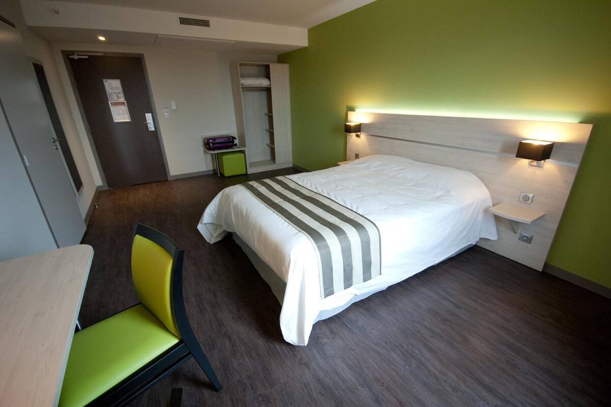 Une chambre d’hôtel comprenant un lit, une chaise, une table, une armoire et une table de chevet avec une lampe.