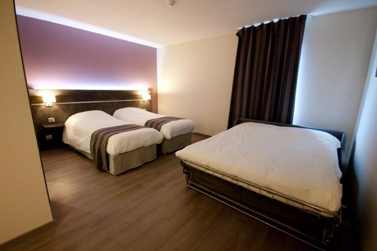 Une chambre d’hôtel avec deux lits simples, un lit plus grand, un plancher en bois et une tête de lit dotée d’un éclairage intégré.