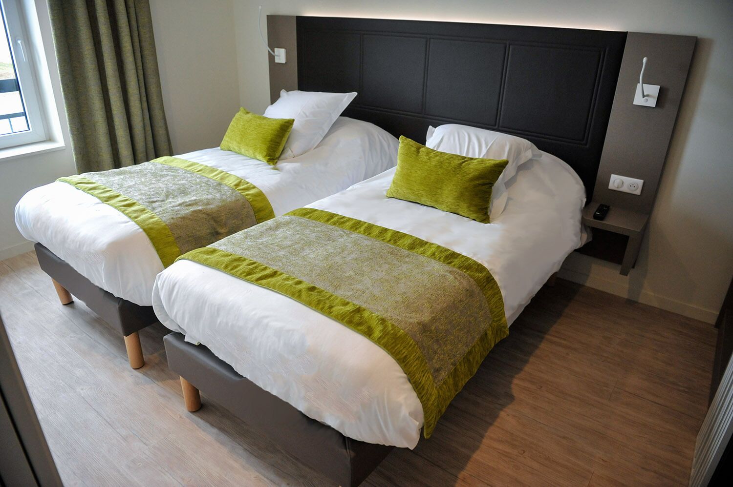 Una habitación de hotel con dos camas, cada una con ropa de cama blanca y detalles verdes, un cabecero oscuro y una mesita de noche pequeña con un control remoto.