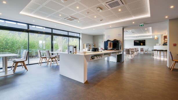 A spacious, modern hotel lobby with a central reception desk, seating area, and large windows offering a view of greenery outside.