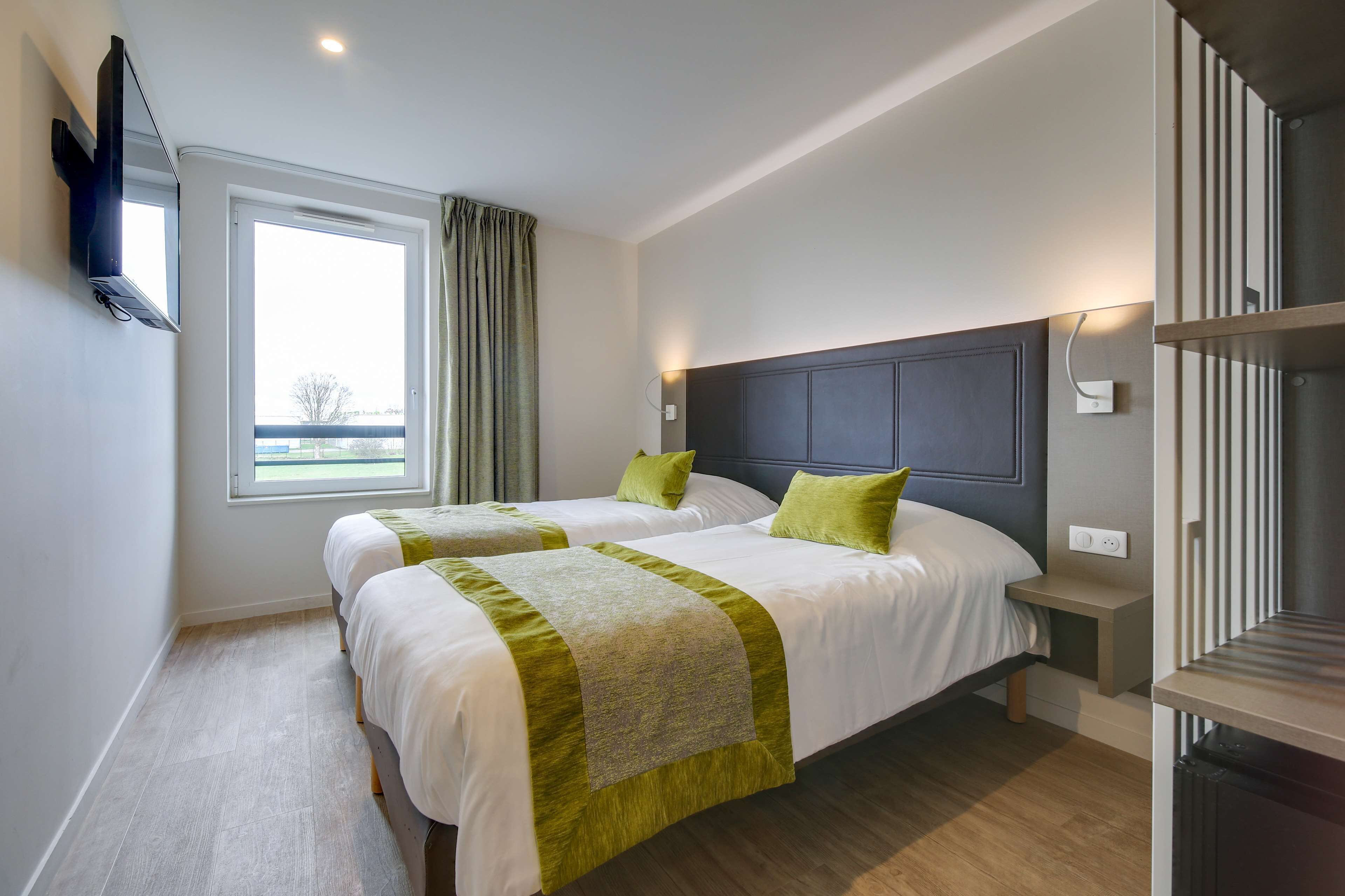 A hotel room with two beds, a large window, a headboard, a bedside table, and a built-in TV.