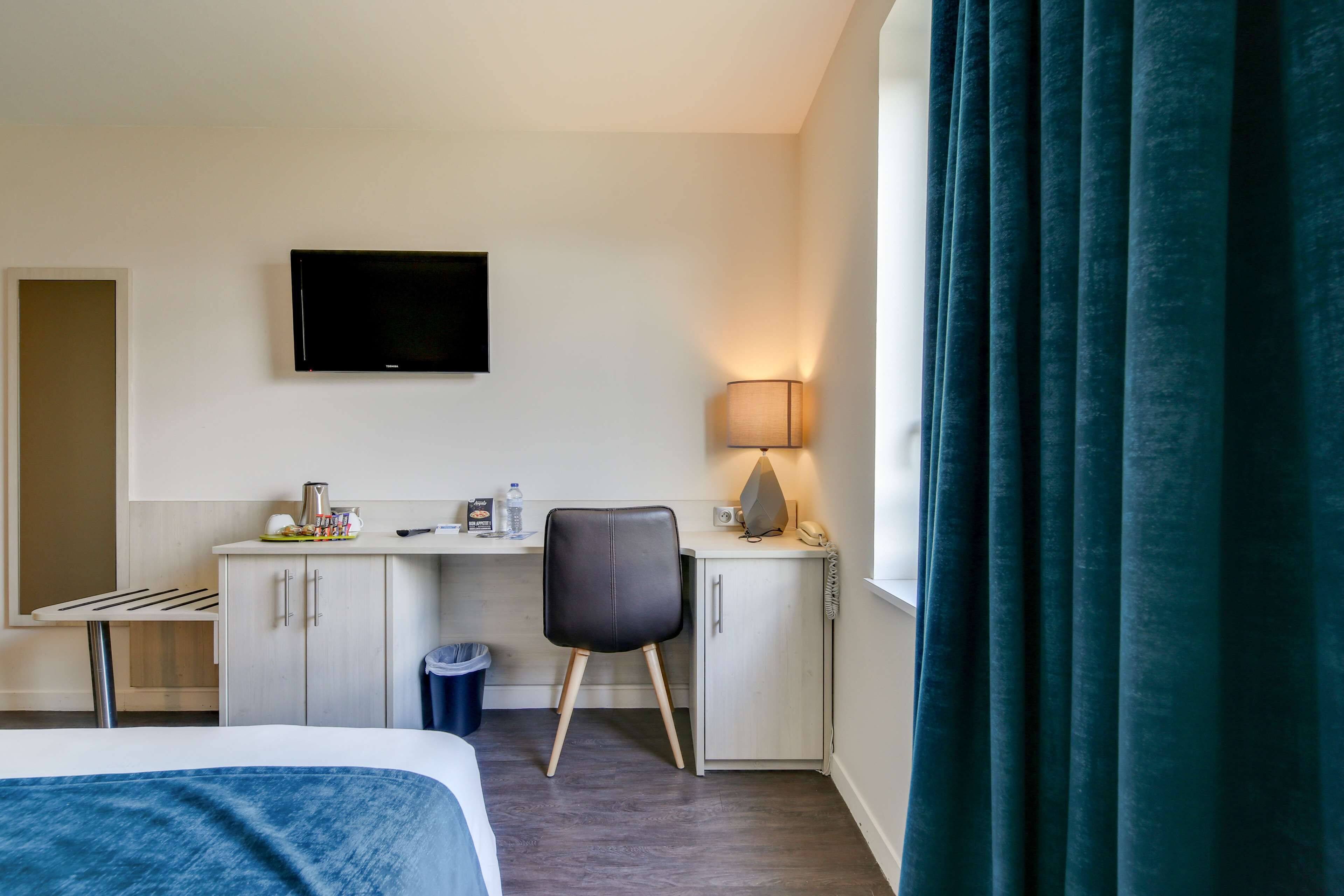 A hotel room with a bed, a desk with a chair, a television, a lamp, and a window with curtains.