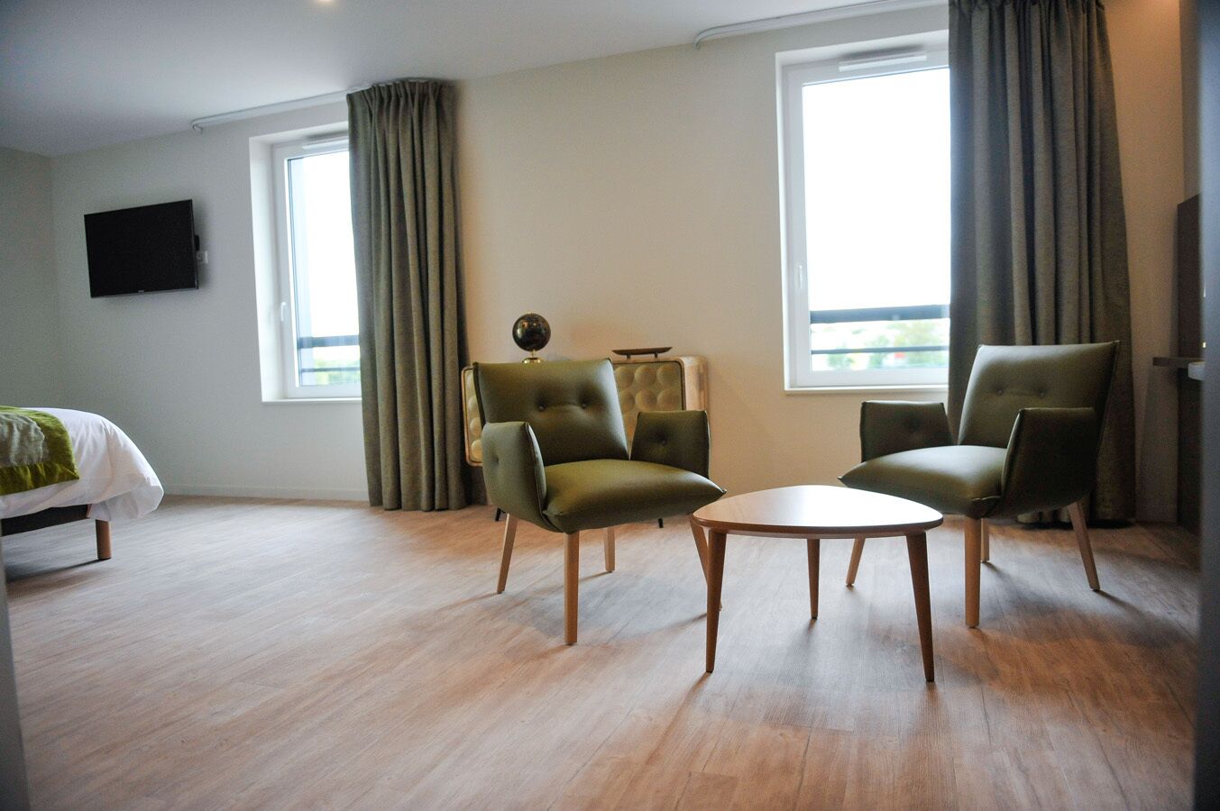 Ein Hotelzimmer mit einem Bett, zwei grünen Sessel, einem runden Couchtisch und einem an der Wand befestigten Flachbildfernseher.