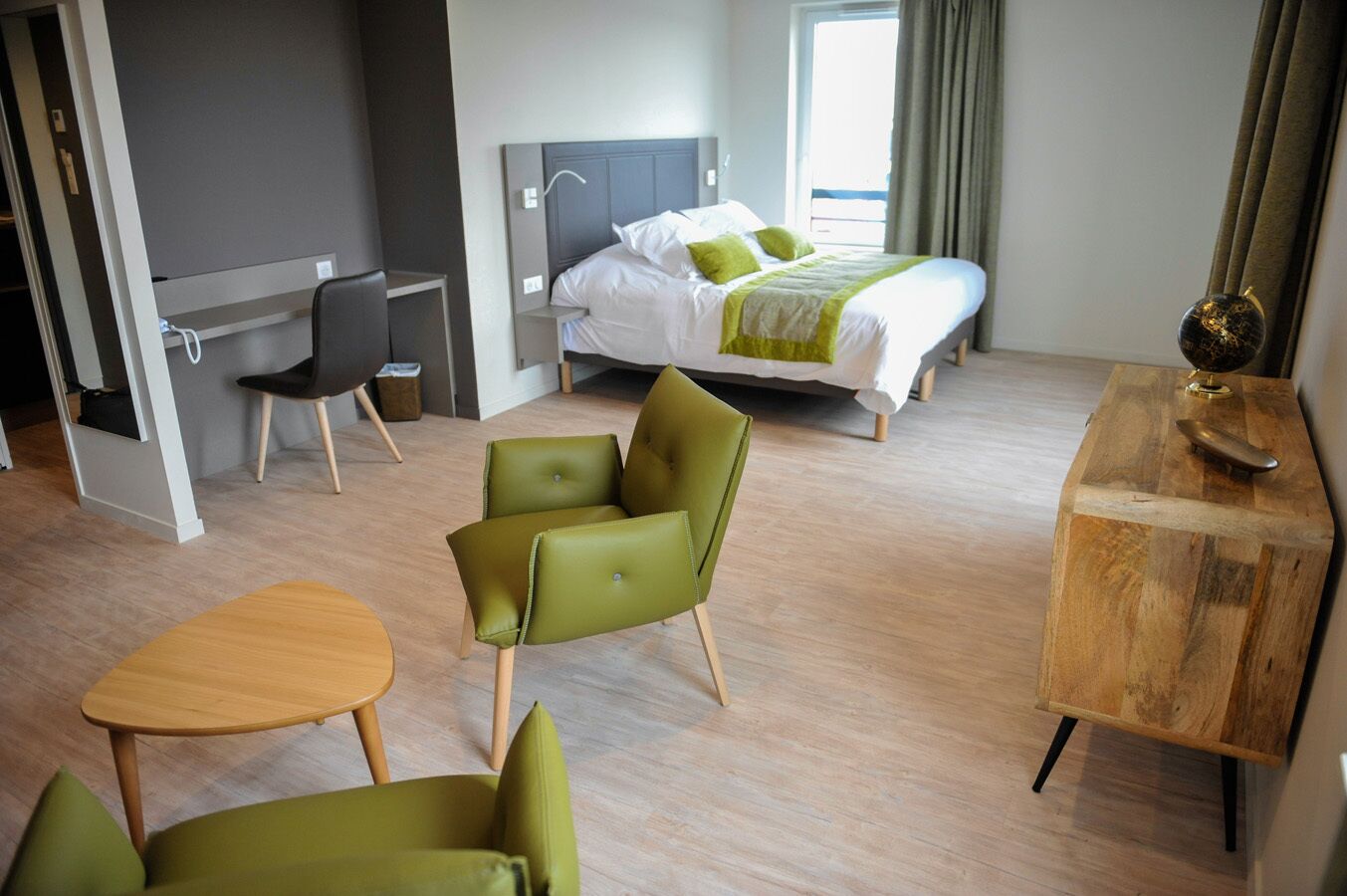 Una habitación de hotel moderna con una cama, un escritorio, dos sillas verdes, una mesa de madera y una mesita de noche de madera.