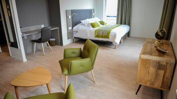 Una habitación de hotel moderna con una cama, un escritorio, dos sillas verdes, una mesa de madera y una mesita de noche de madera.
