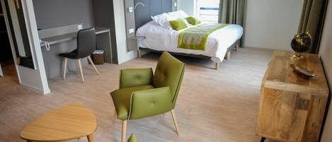 A modern hotel room with a bed, a desk, two green chairs, a wooden table, and a wooden side table.