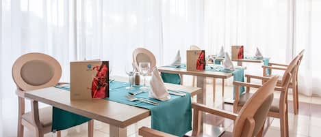 Un espace repas avec des tables et des chaises en bois, des nappes de couleur turquoise et une grande fenêtre ornée de rideaux légers.