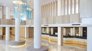 Een moderne lobby met een receptiebalie, grote zuilen en een hoog plafond met hangende lampen.