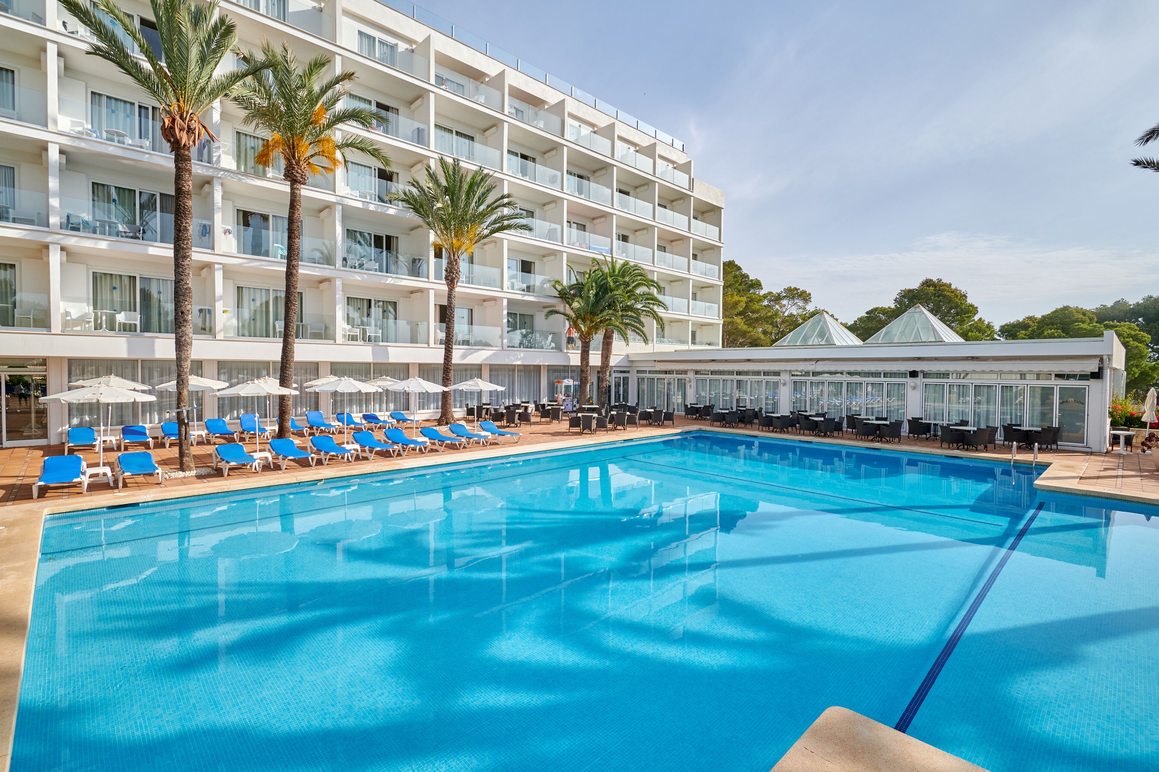 A hotel pool area with multiple lounge chairs and umbrellas, a large swimming pool, and a building with balconies and a covered area.