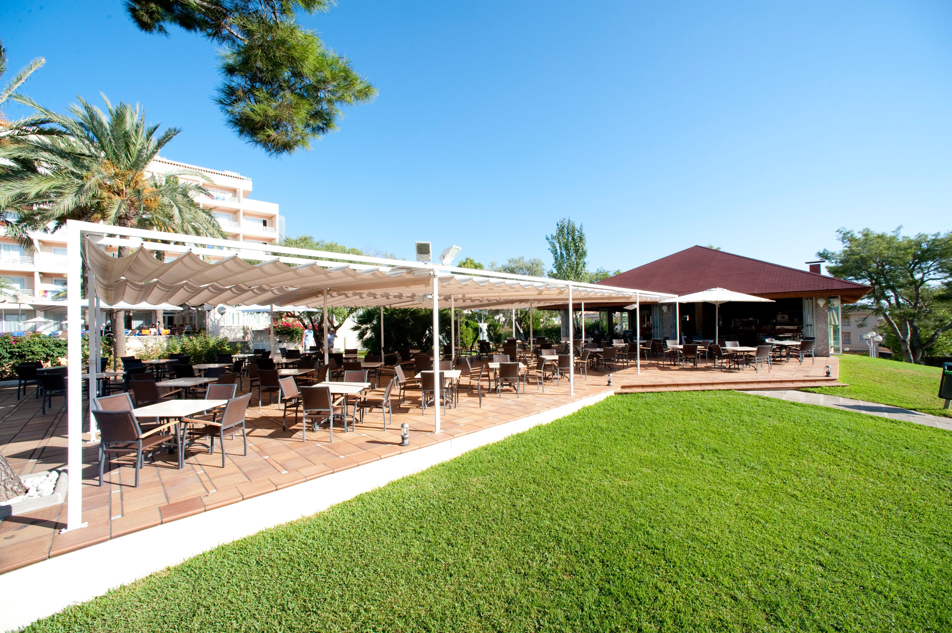 An outdoor patio area with tables and chairs, a covered seating area, and a building with a red roof in the background.