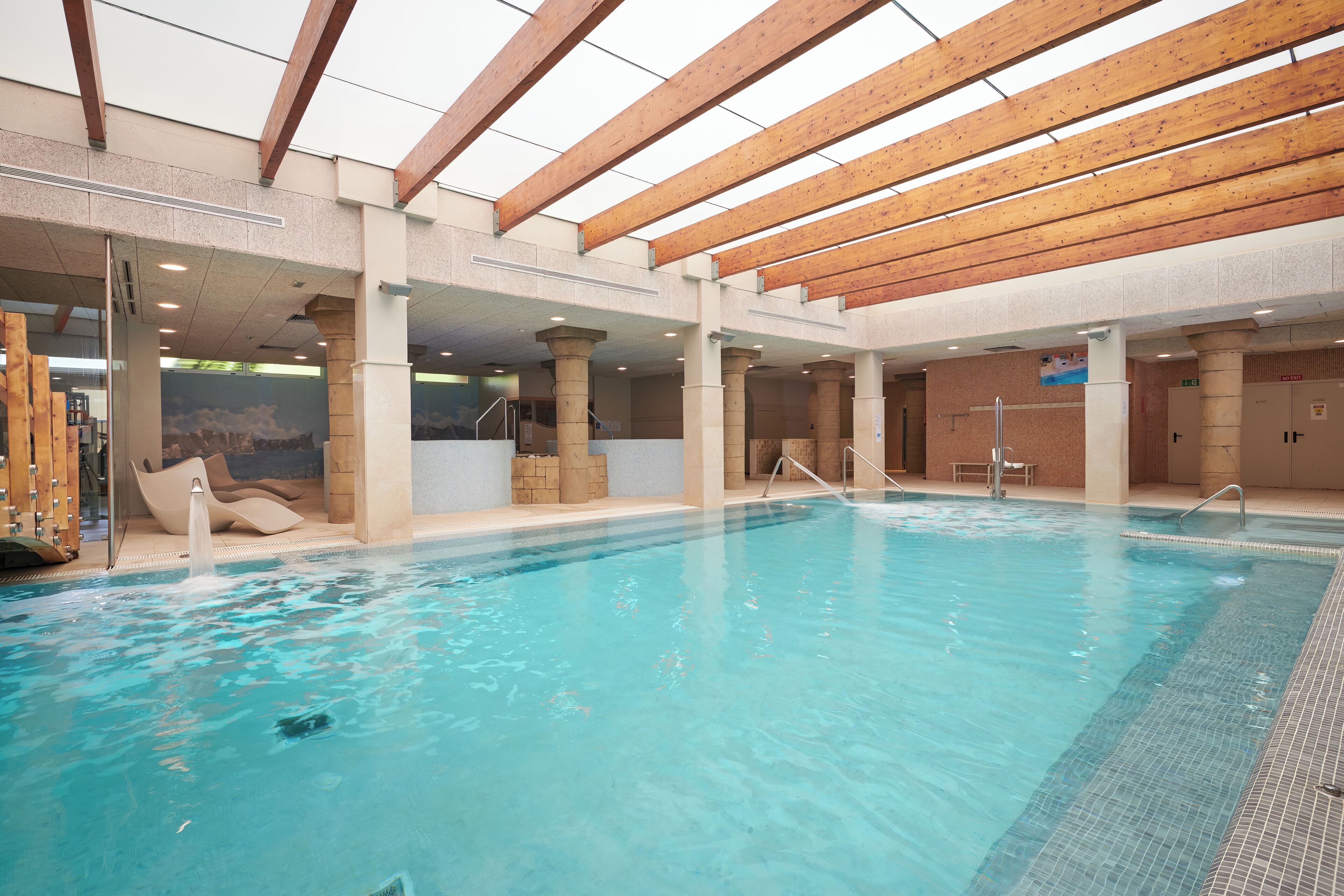 An indoor swimming pool with clear water, wooden beams, and a tiled floor.