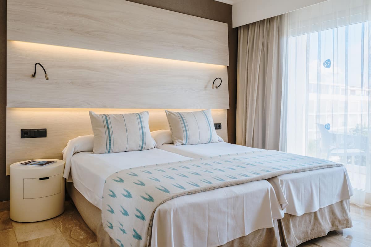 Ein modernes Hotelzimmer mit zwei Betten, einem großen Fenster mit Vorhängen und einem Holzkopfteil.