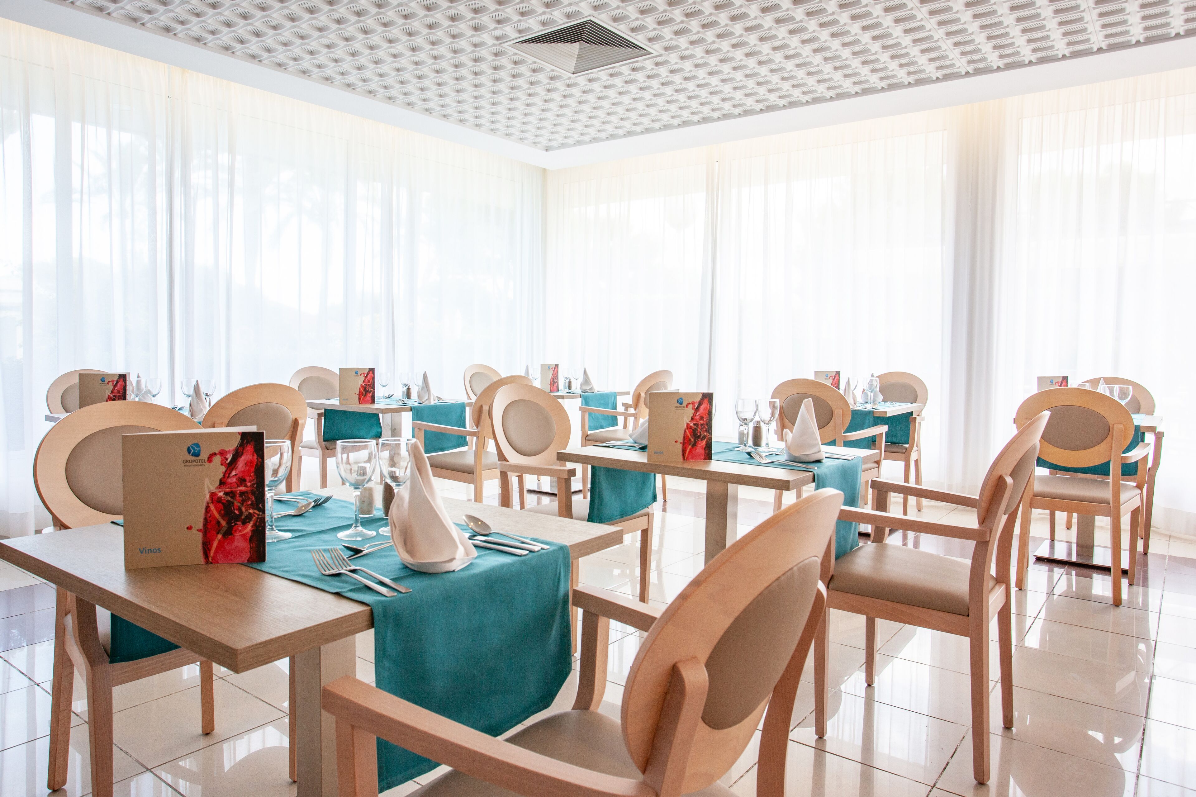 Zona pranzo con tavoli apparecchiati per quattro, sedie di legno e una stanza luminosa e ben illuminata dalla luce naturale, con ampie finestre.